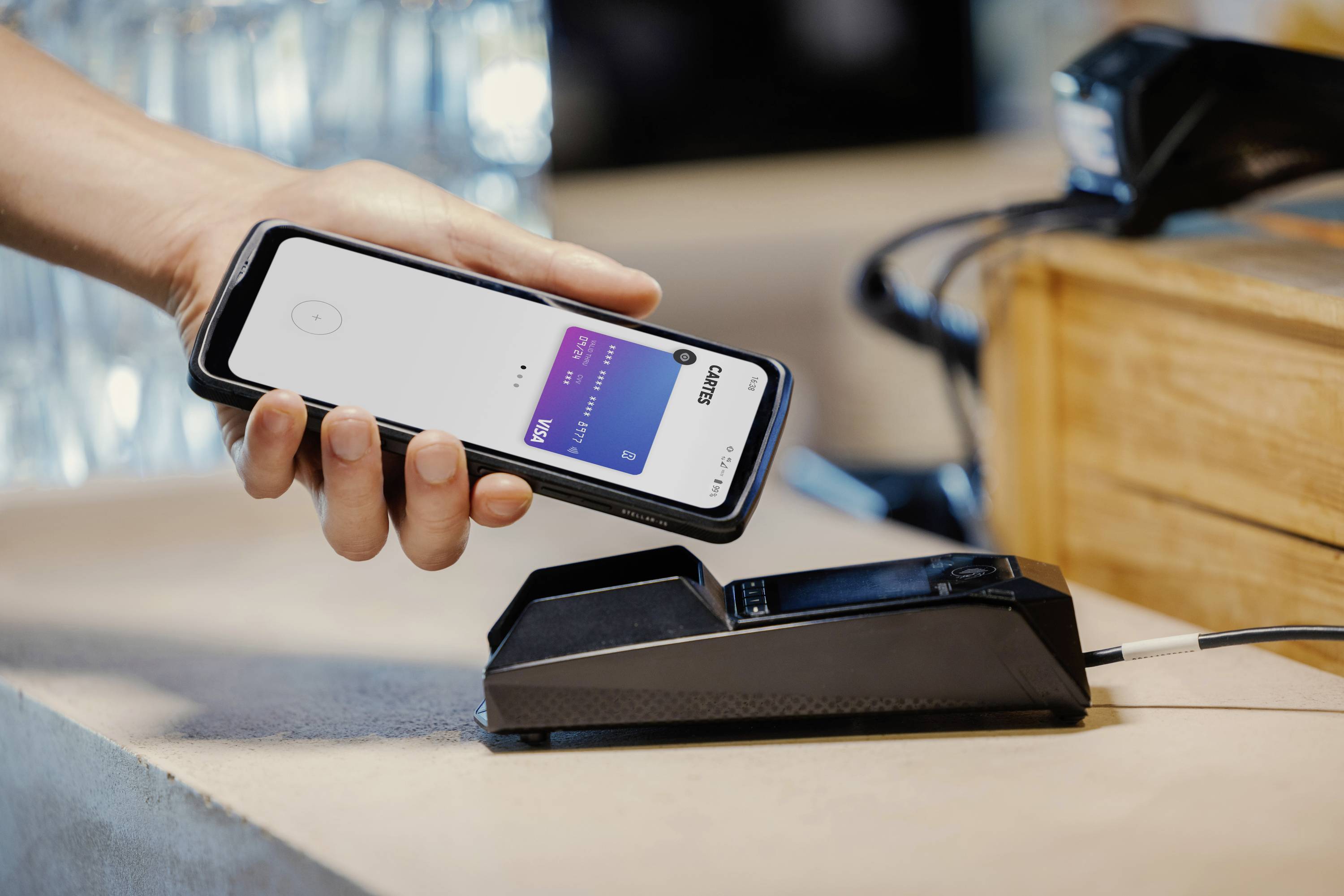 A hand is holding a smartphone with an open payment app over a card terminal used for contactless payment.