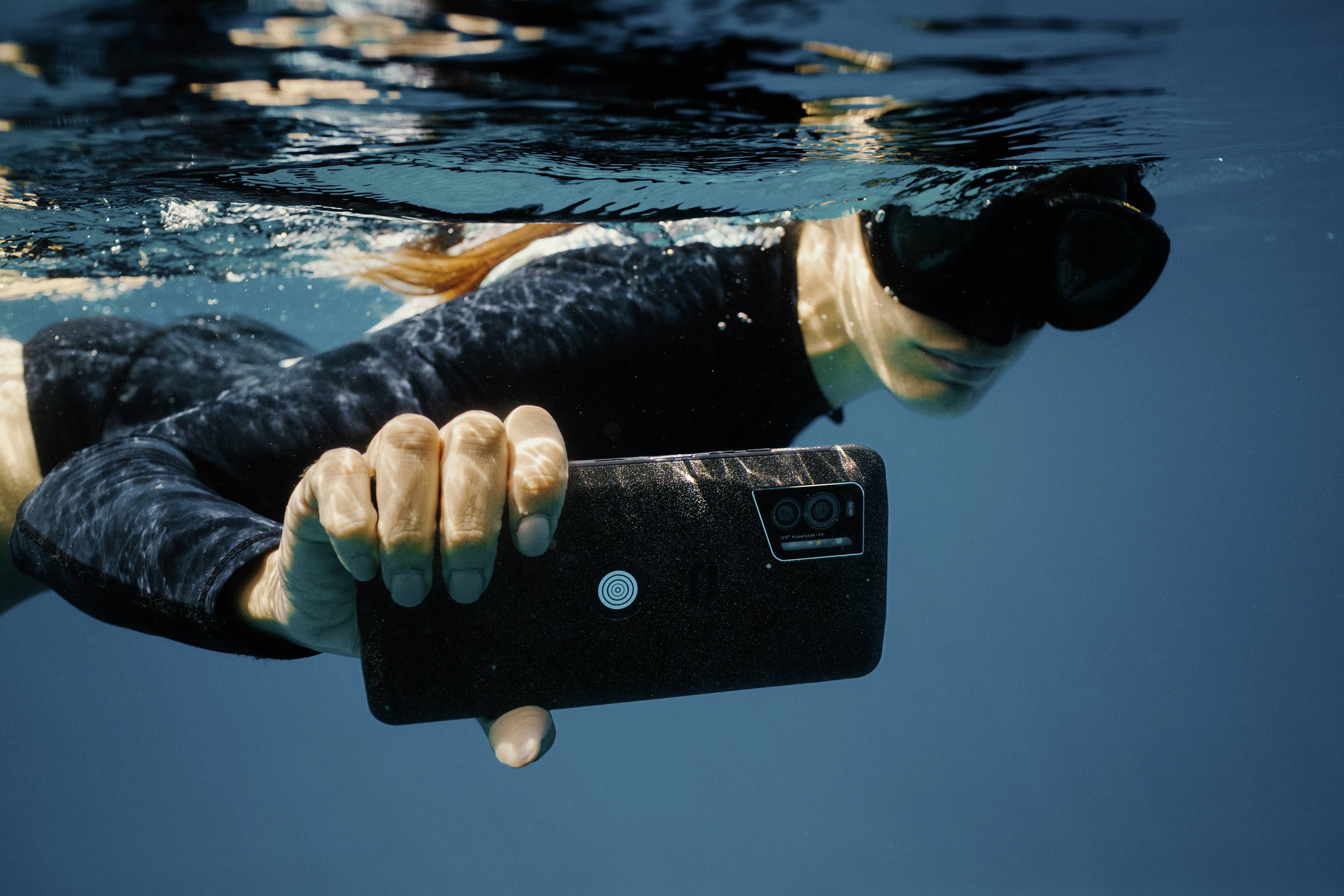 A person is diving underwater, holding a smartphone and wearing diving goggles.