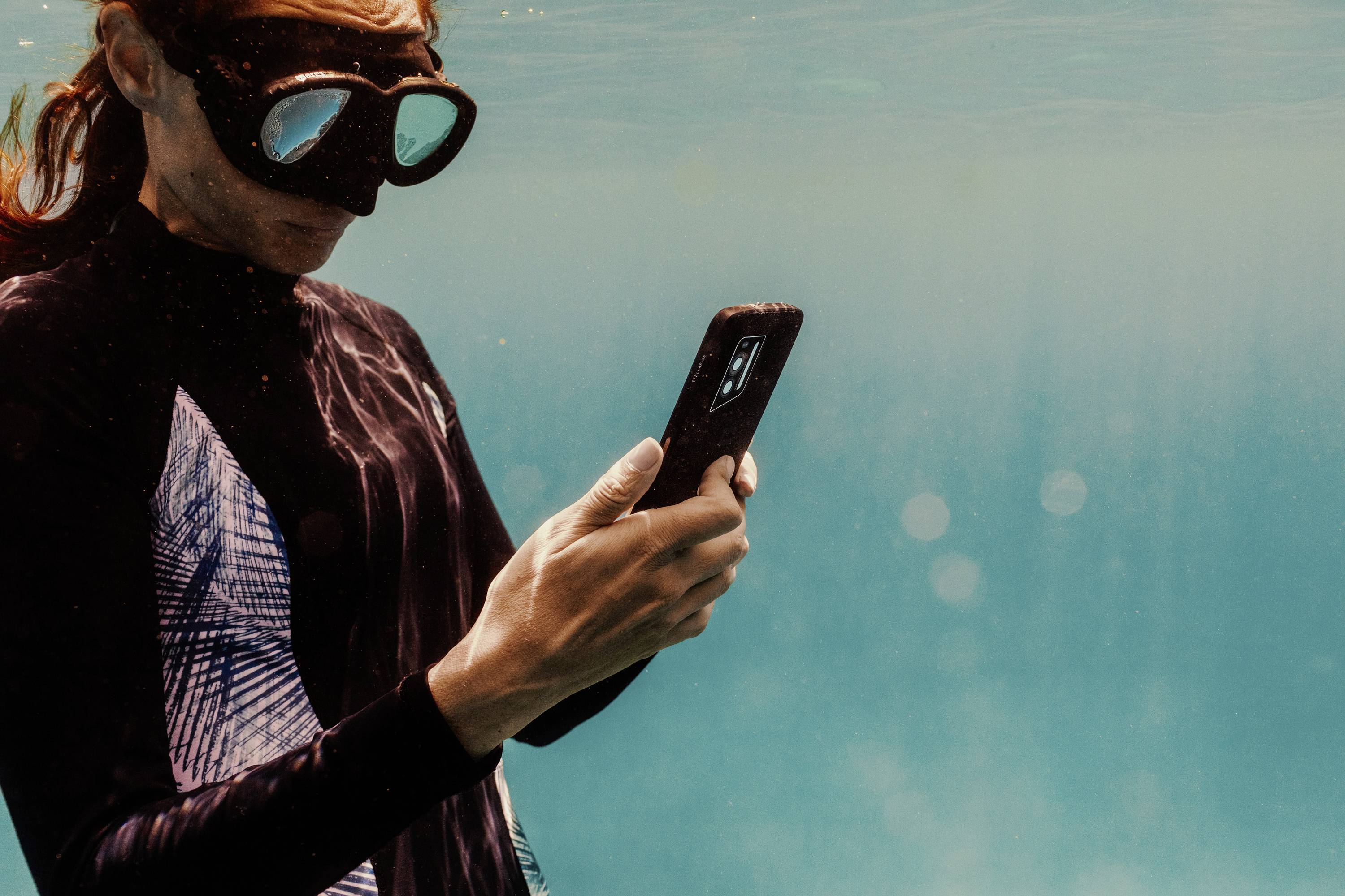 A person is wearing a diving mask and using a smartphone underwater. The environment is blue-green, suggesting a sea or swimming pool.