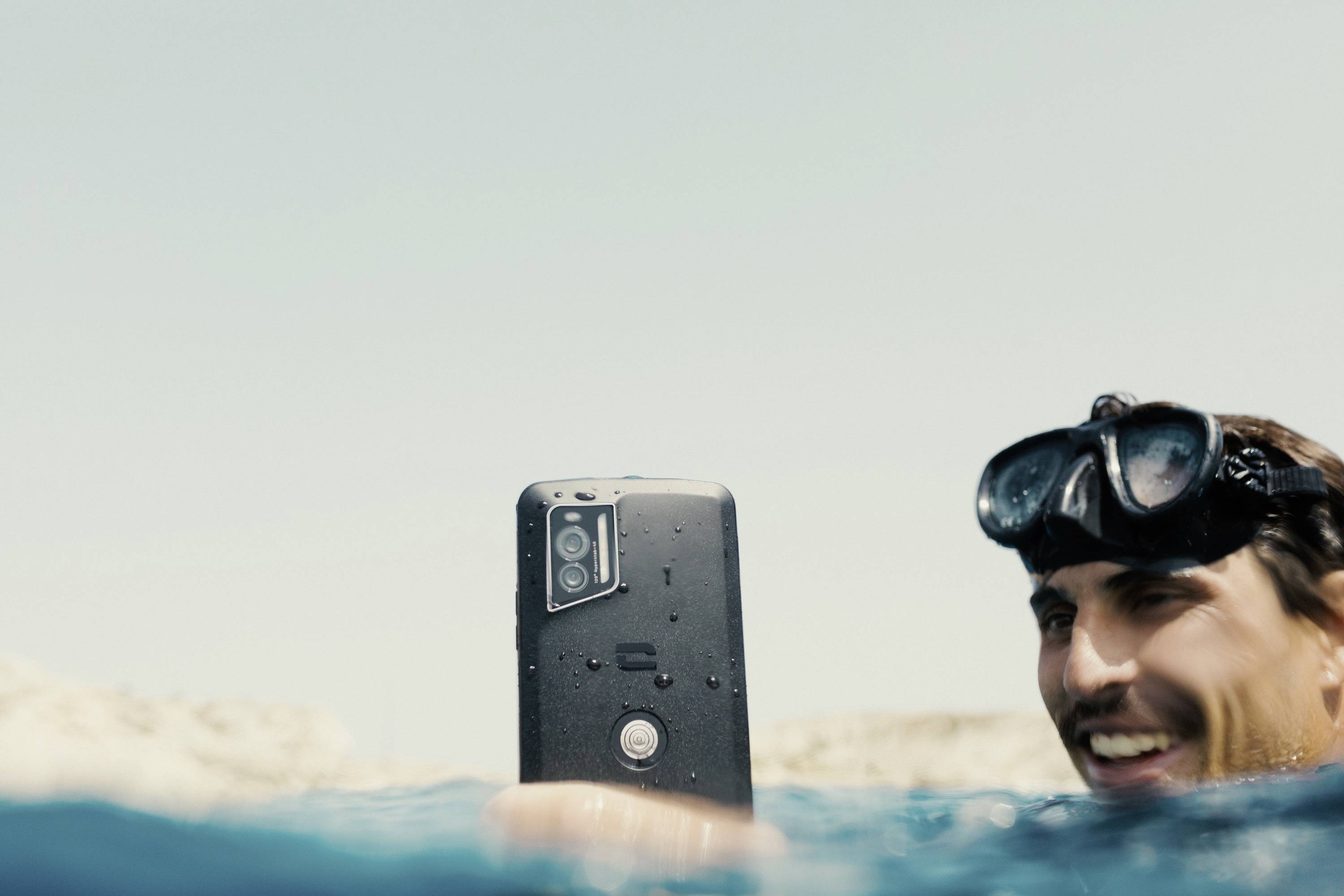Man wearing diving goggles holds waterproof smartphone above water surface, smiling, bright sky in background.