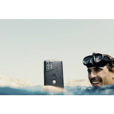 Man wearing diving goggles holds waterproof smartphone above water surface, smiling, bright sky in background.
