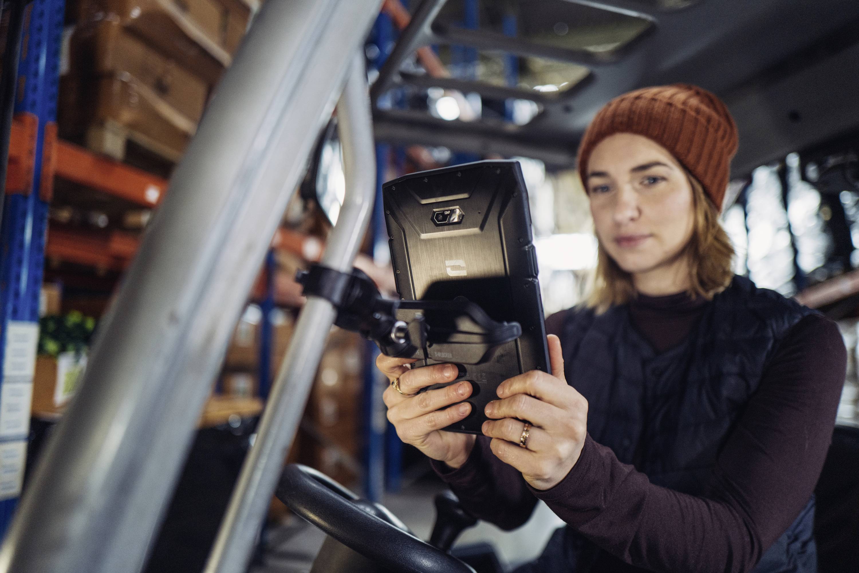 A person wearing a cap is looking at an electronic device in a warehouse with shelves in the background.