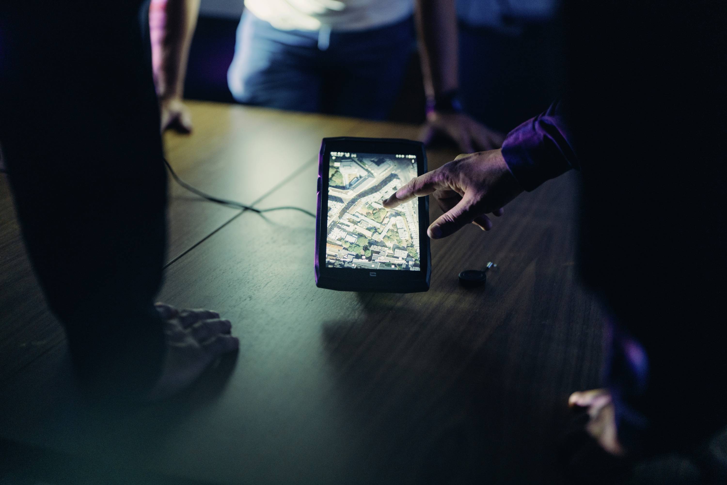 A person is pointing at a tablet displaying a map view. Several people are looking on, concentrating intently on the map. The atmosphere is that of a conference setting.