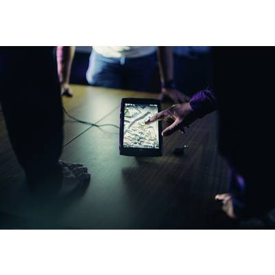 A person is pointing at a tablet displaying a map view. Several people are looking on, concentrating intently on the map. The atmosphere is that of a conference setting.