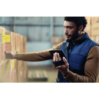 A man is checking stock levels in a warehouse using a tablet. He is standing in front of shelves stacked with boxes.
