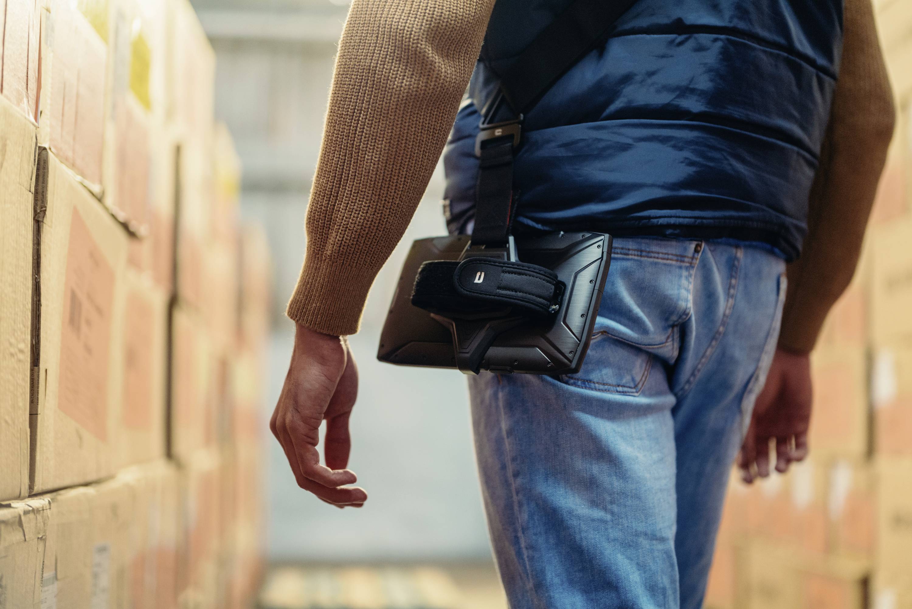 A person wearing a jacket is carrying an electronic device on their hip in a warehouse full of cardboard boxes.