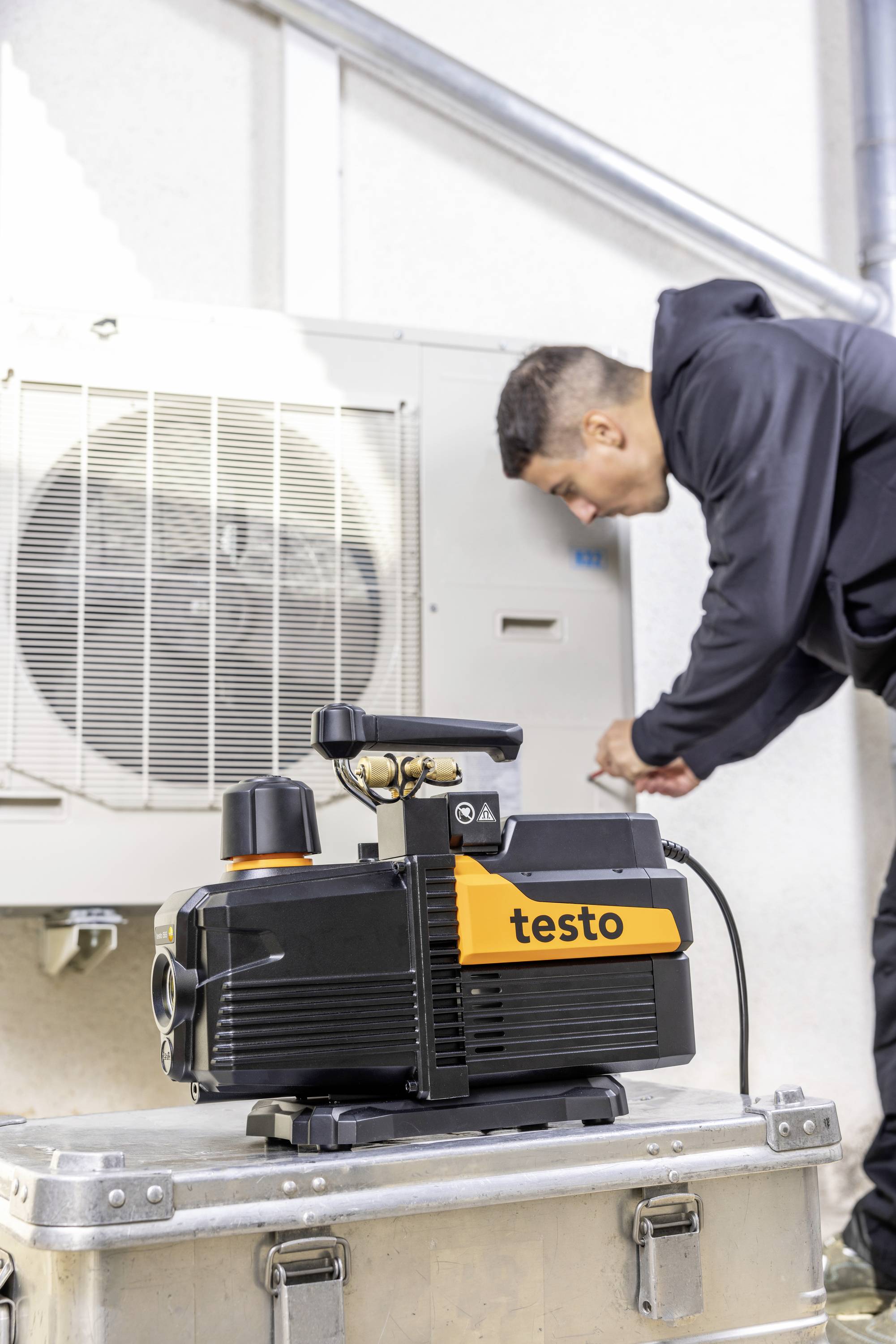 A technician is working on an air conditioning unit. In the foreground, a testing device sits on a case.