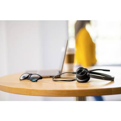 A headset, a pair of glasses, and a laptop on a table. In the background, a person is blurrily visible standing by a window.