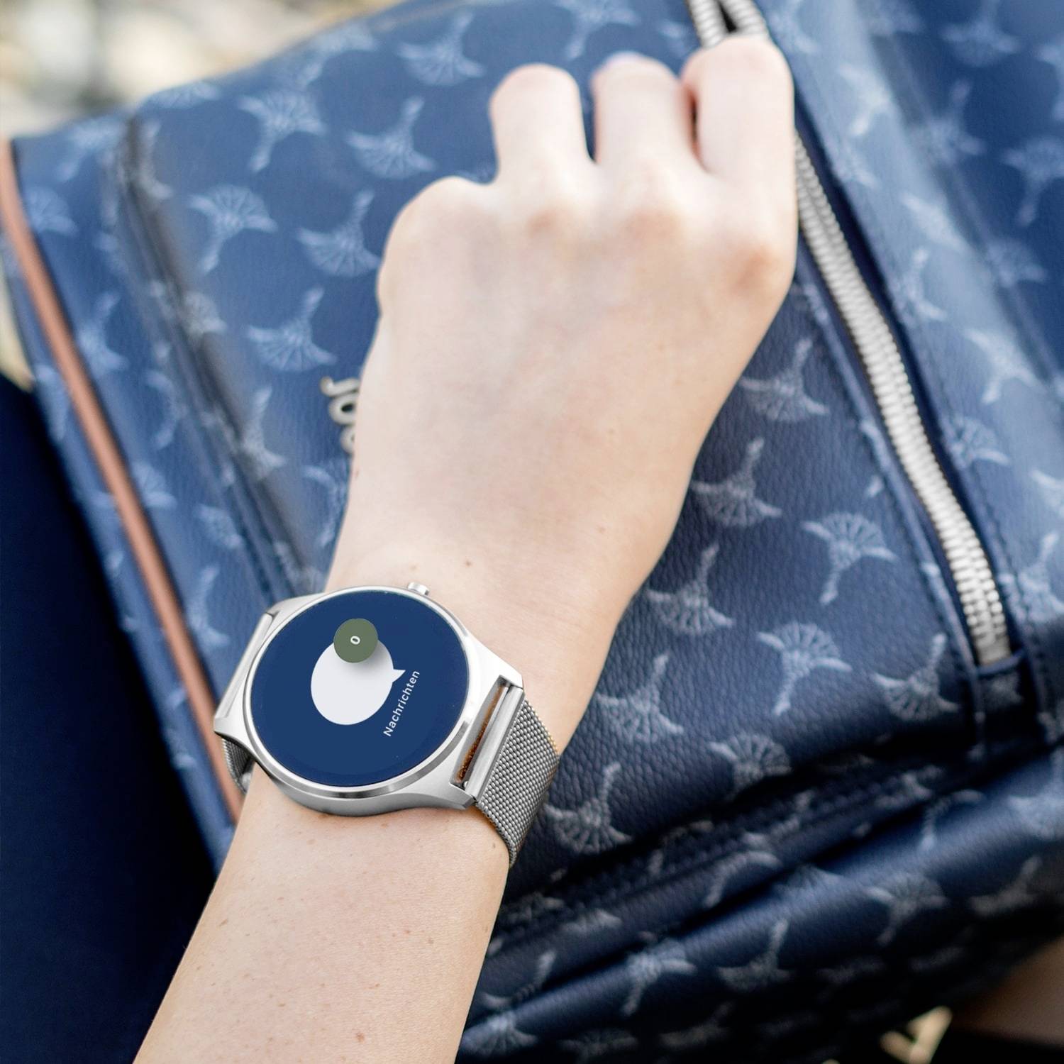 A hand is holding a smartwatch with a blue dial displaying a notification. Part of a rucksack is visible in the background.