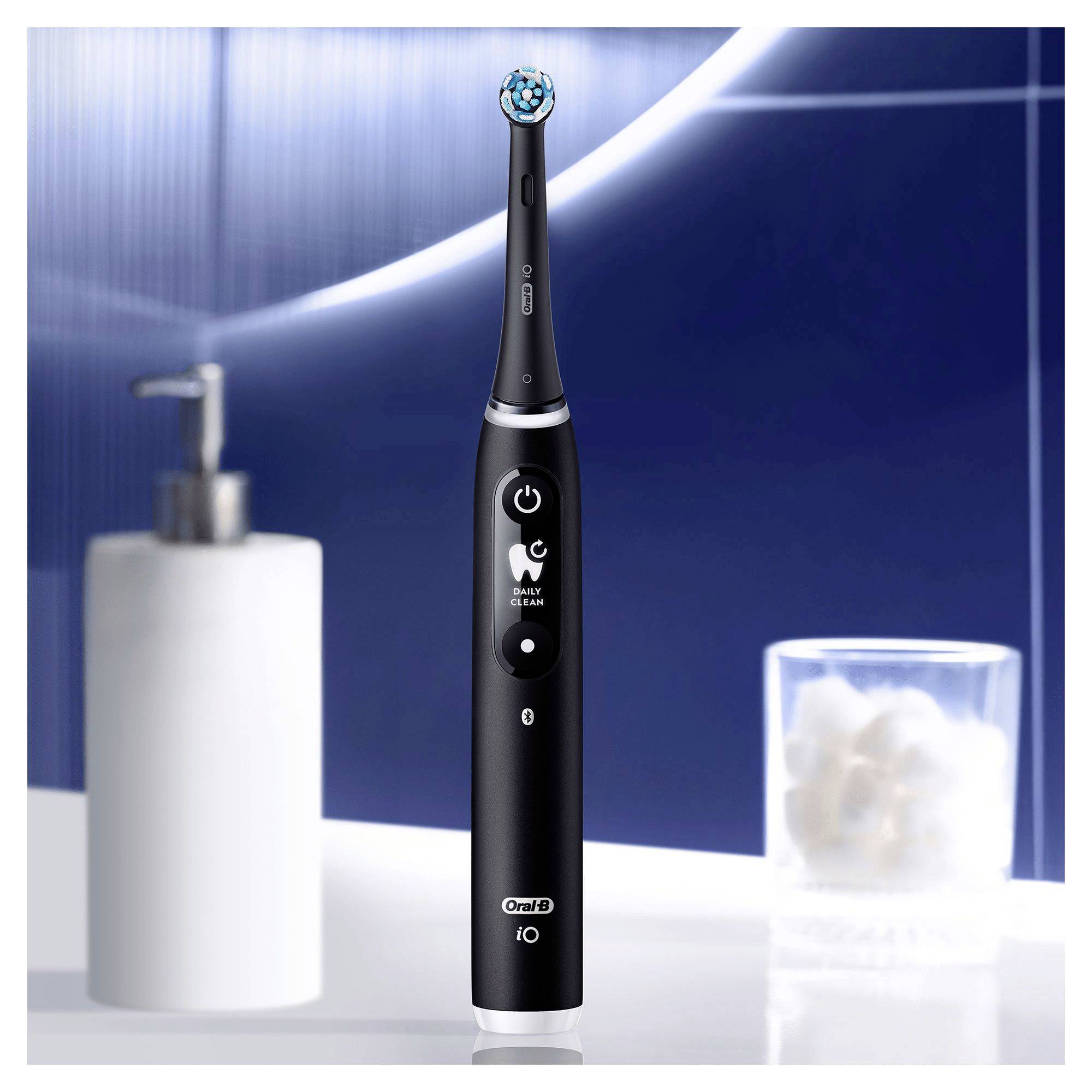 Black electric toothbrush in the bathroom. A soap dispenser on the left, a glass with cotton wool balls on the right. Background blurred.