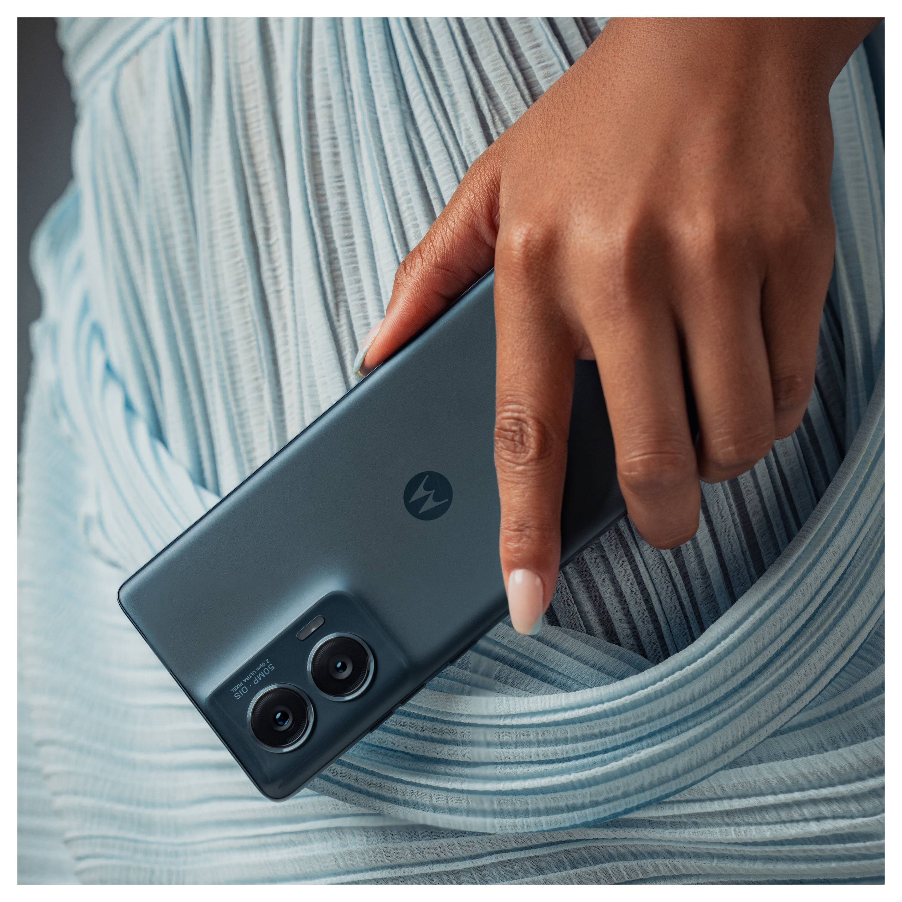 A hand holding a smartphone with dual cameras, resting against a pleated, light blue fabric, creating a stylish and modern aesthetic.