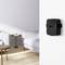 A black Apple TV box is mounted on the white wall of a modern living room. In the background, a table and a sofa are visible.