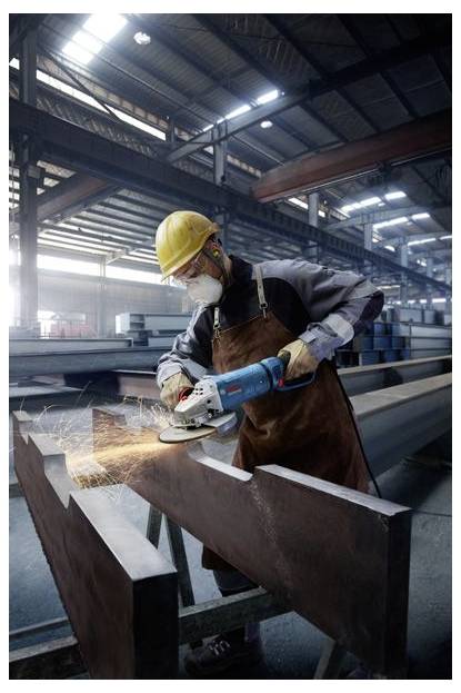 A factory worker in safety gear grinds a metal beam with a power tool, creating sparks, in a spacious, industrial warehouse setting.