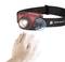 A hand activates a red head torch using gesture control. The lamp shines brightly and is mounted on a black headband.