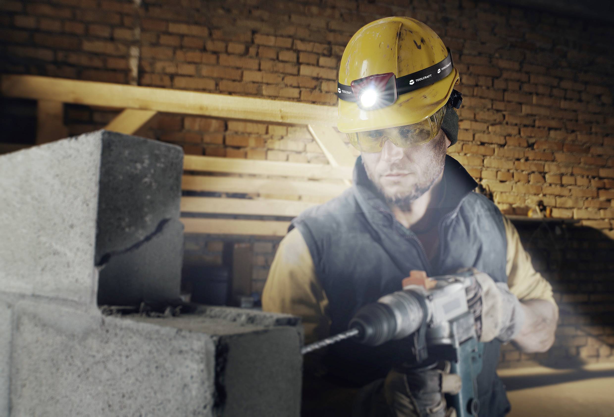 A construction worker in protective clothing and a hard hat is using a drill to bore a hole into a cement block. A brick wall is visible in the background.
