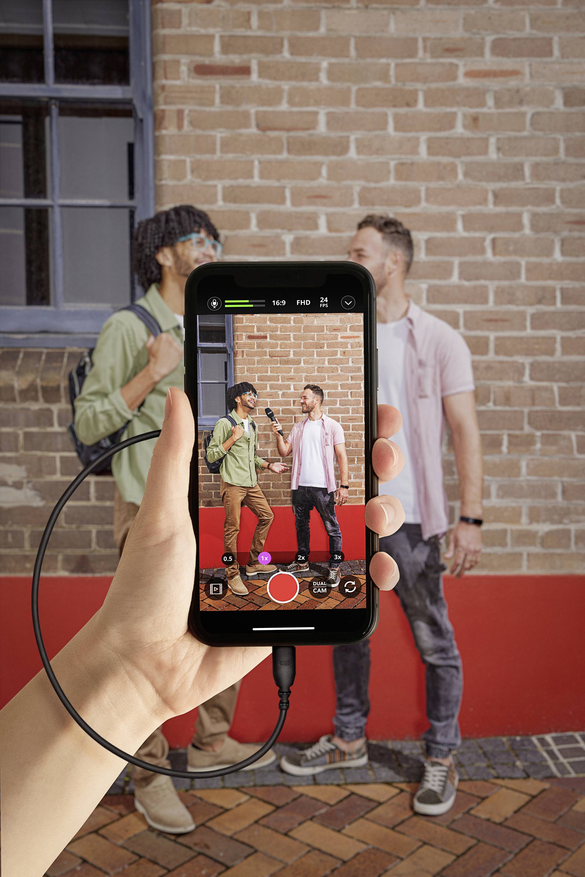 Two men are conversing in front of a brick wall. In the foreground, a smartphone is filming the scene, held in a person's hand.
