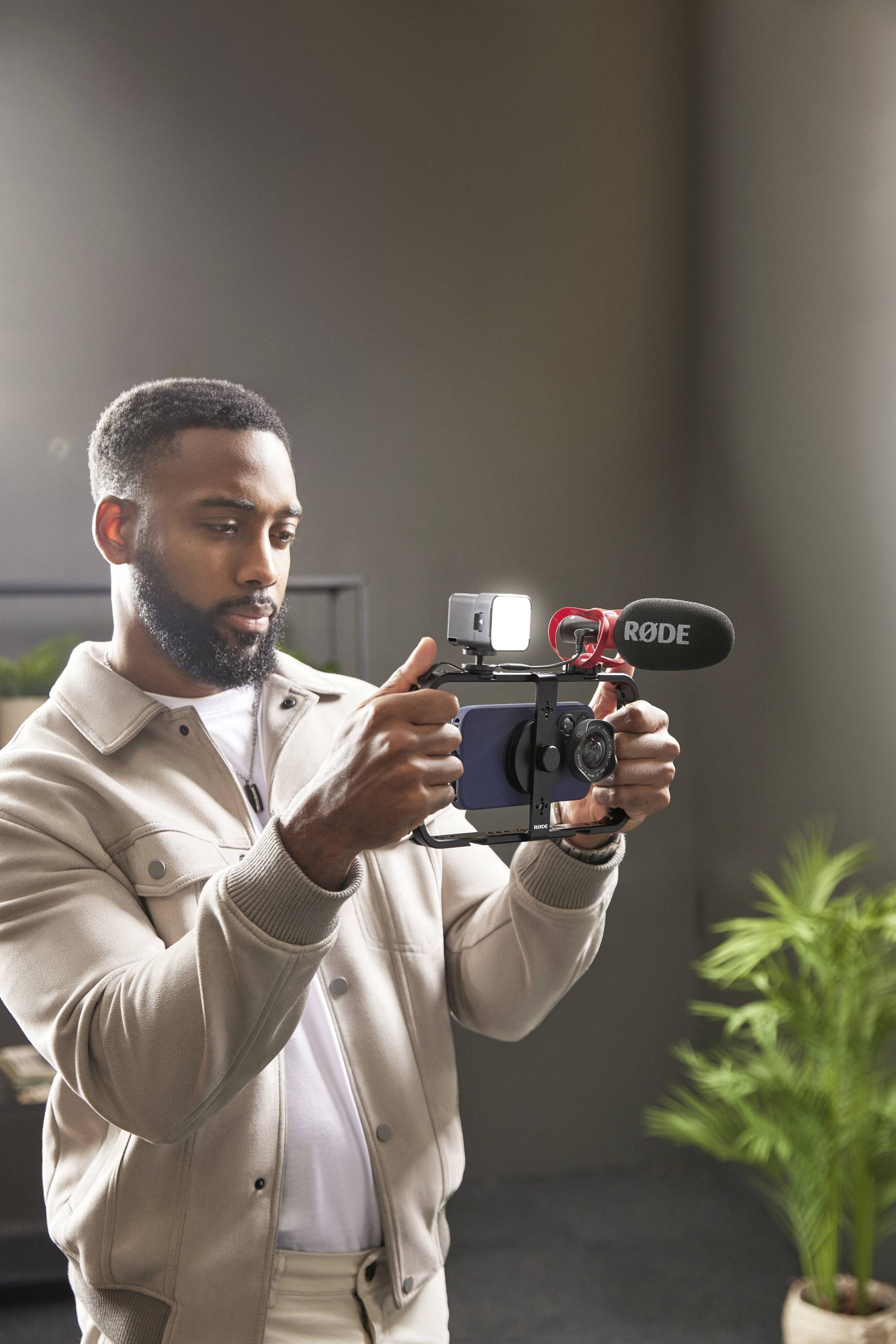 A man is standing in a room with plants, holding a camera with a microphone and lighting. He appears to be recording a video.
