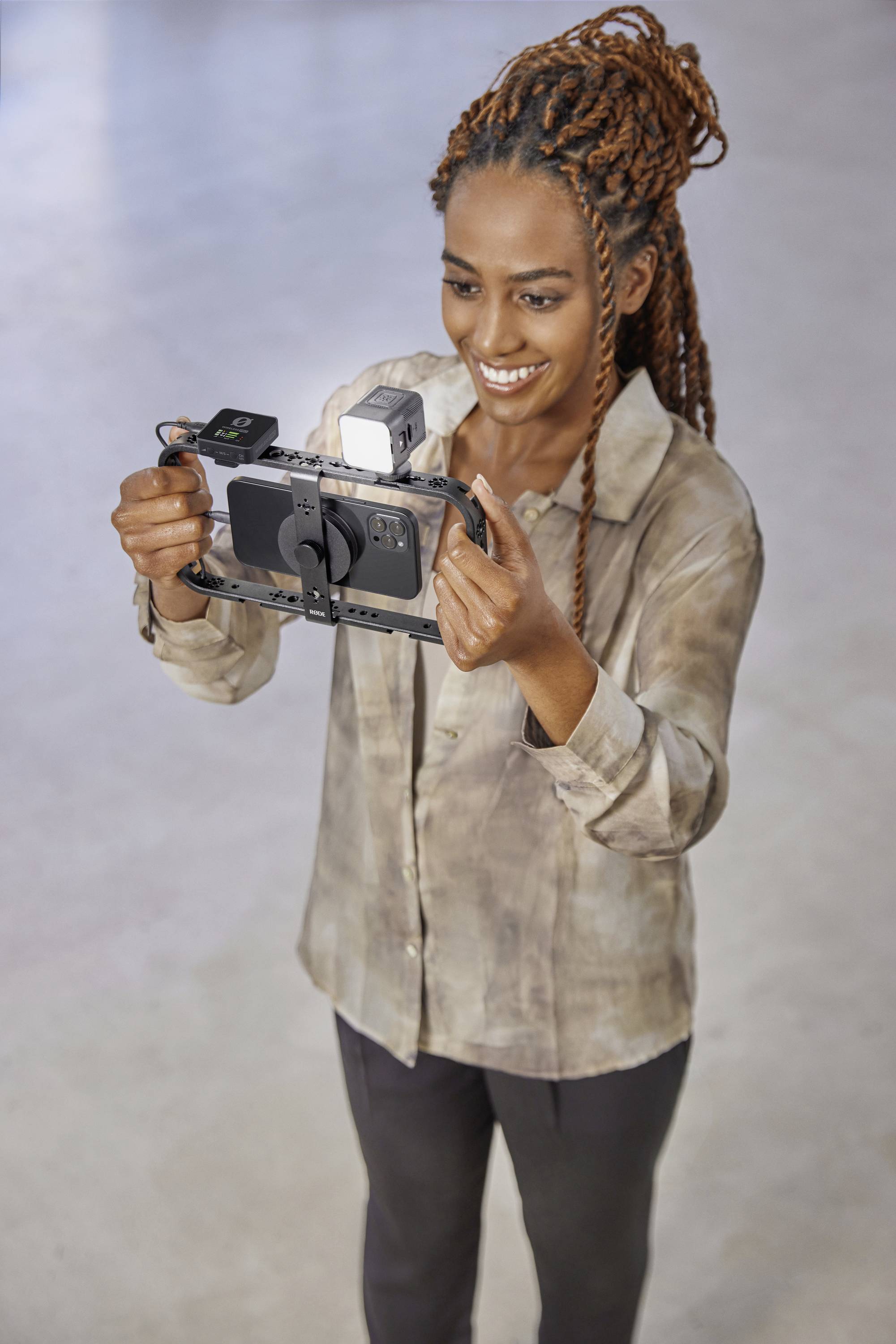 A woman is holding a mobile phone with camera equipment and smiling. She is standing in a bright room.