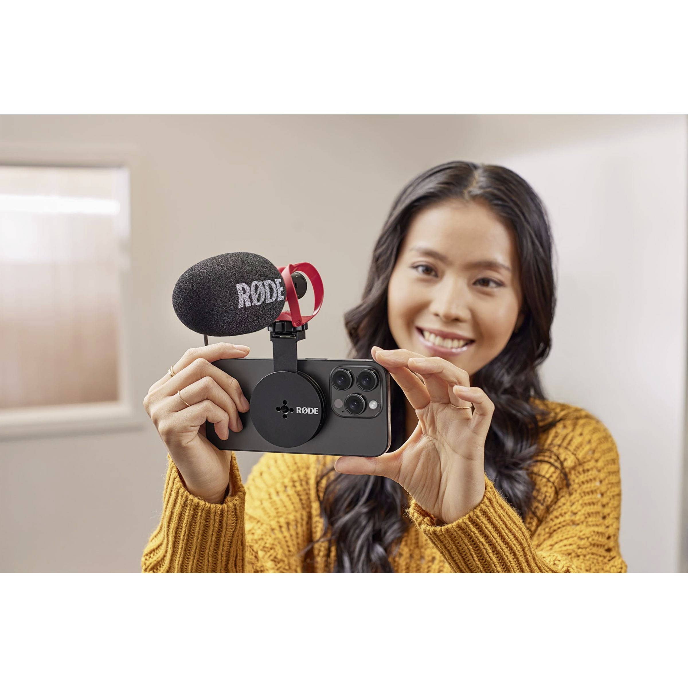 A person is holding a smartphone with an external microphone attached, smiling and appears to be taking a selfie.