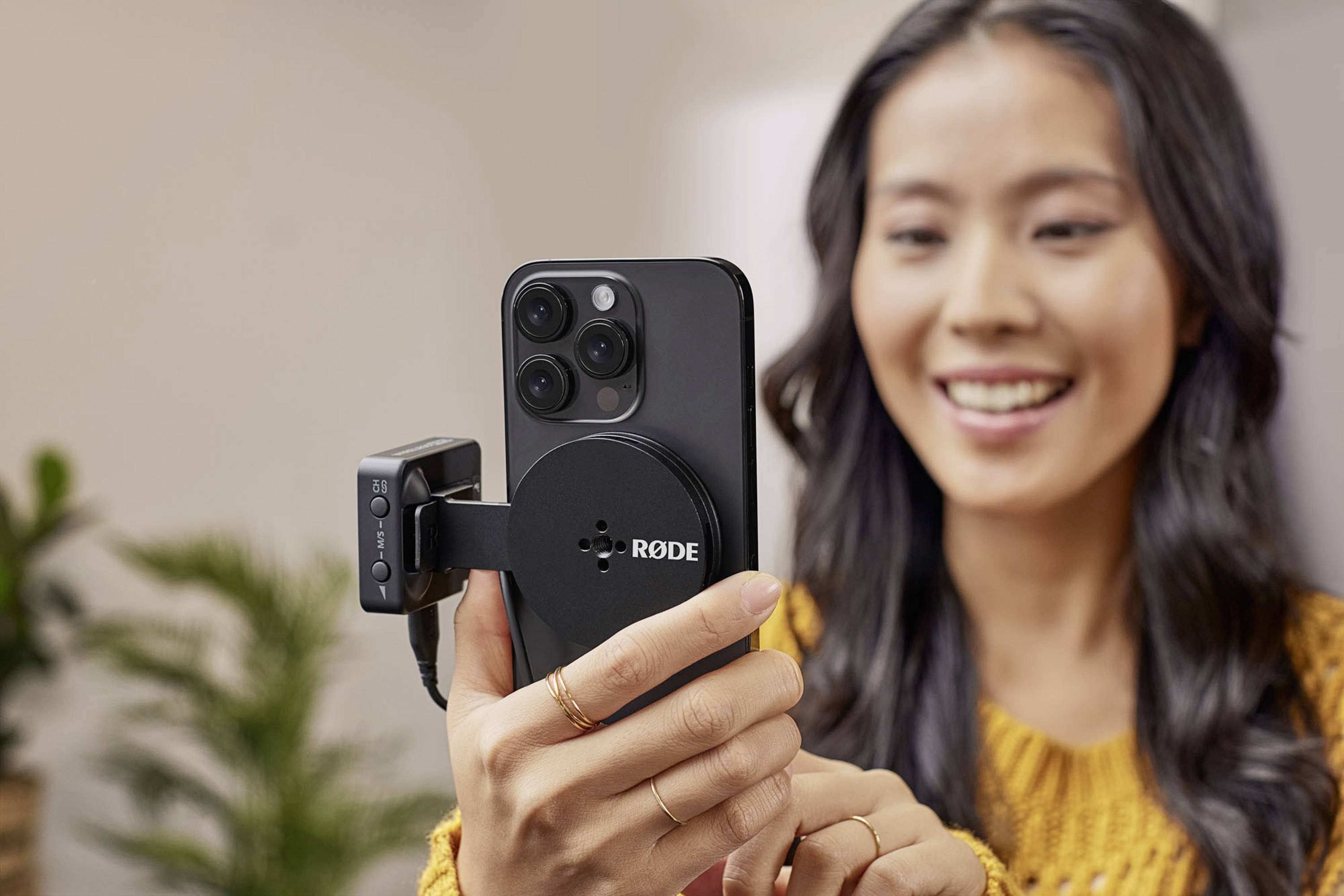 A smiling woman is holding a smartphone with a microphone connected, likely during a video recording.