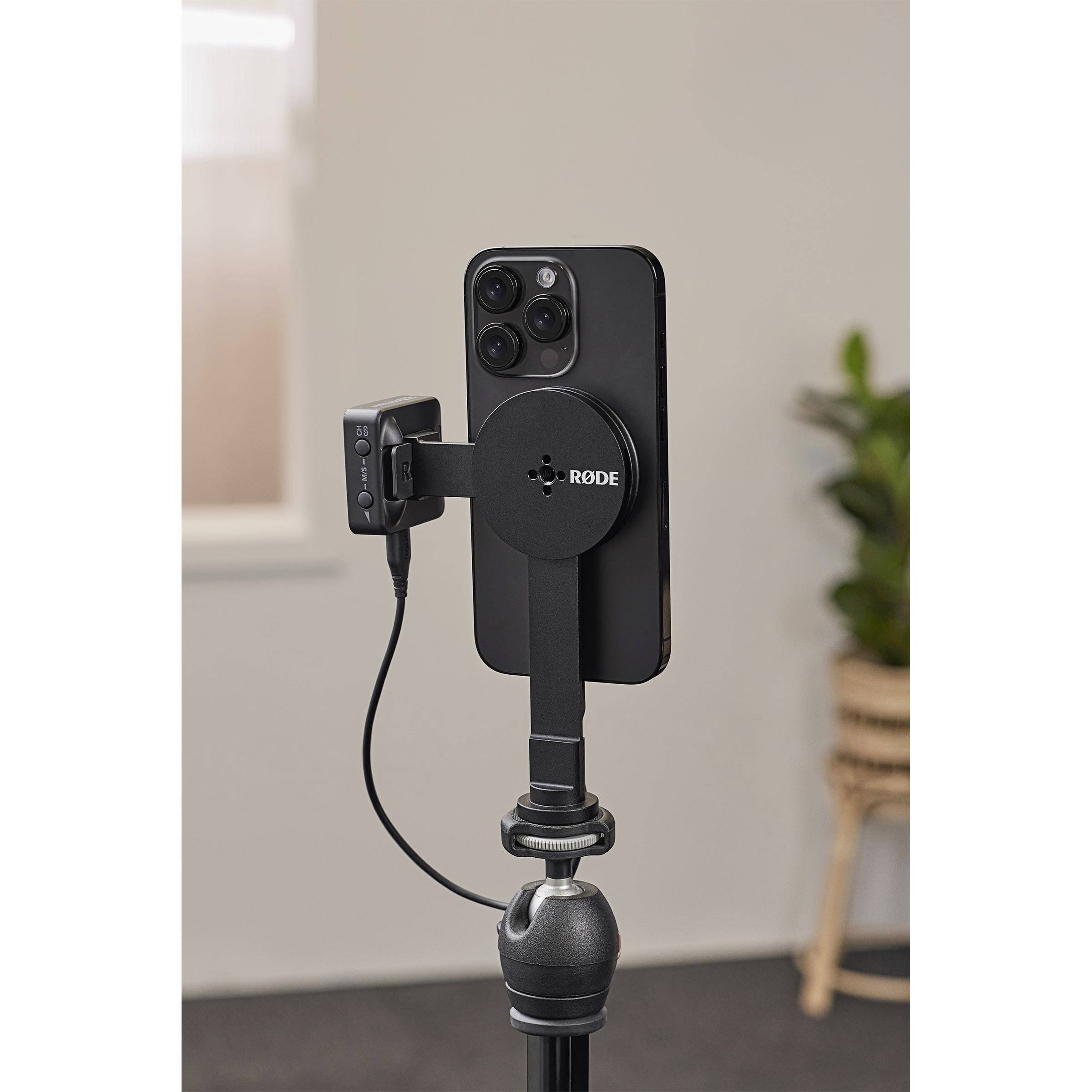 A smartphone is mounted on a tripod, with a RODE microphone connected. A plant is visible in the background.