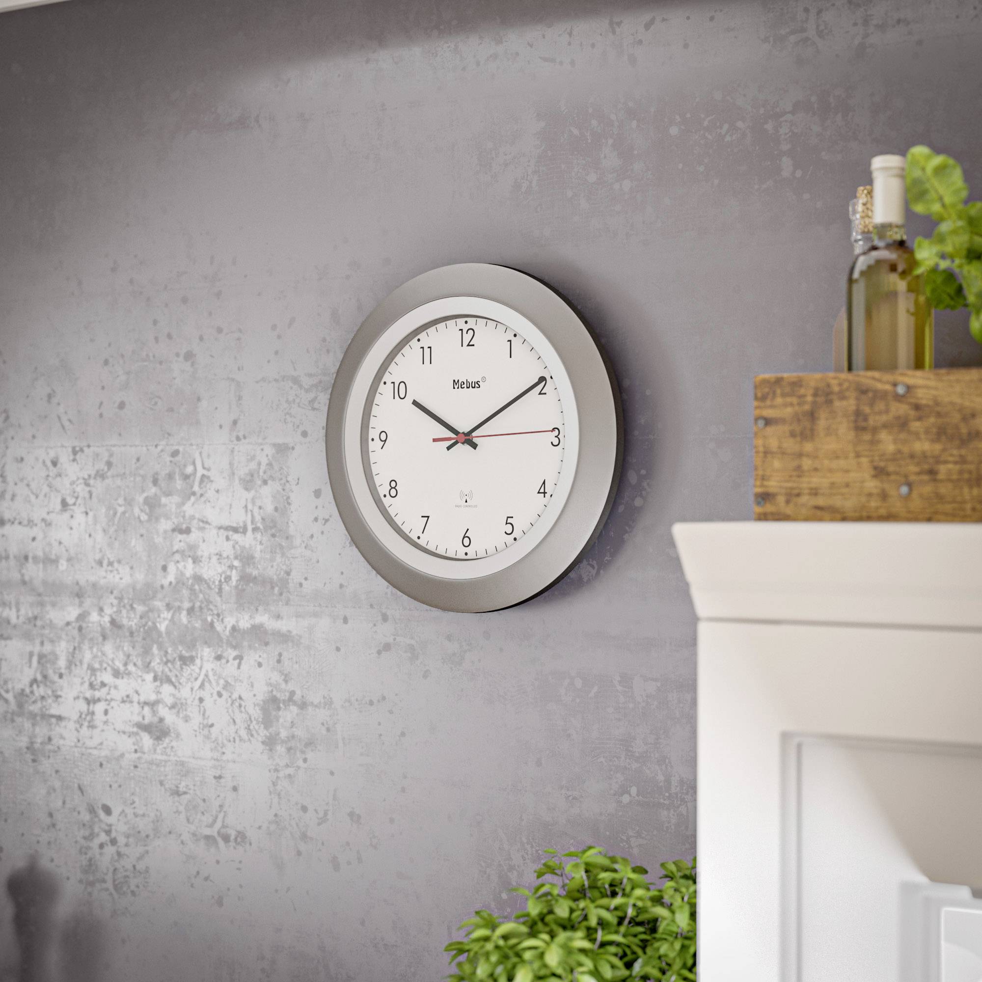 A round wall clock with white dial faces and black hands hangs on a grey wall. A plant is visible in the bottom left.