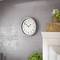 A round wall clock with a black frame shows 10:10. It hangs on a grey wall next to a shelf with plants.