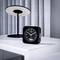 A black alarm clock with a white dial and digital temperature display sits on a bedside table, next to a modern lamp.