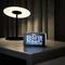 A digital alarm clock on a table shows the time as 13:00, date 16th April, day Thursday, and temperature 23.6°C. A table lamp stands in the background.