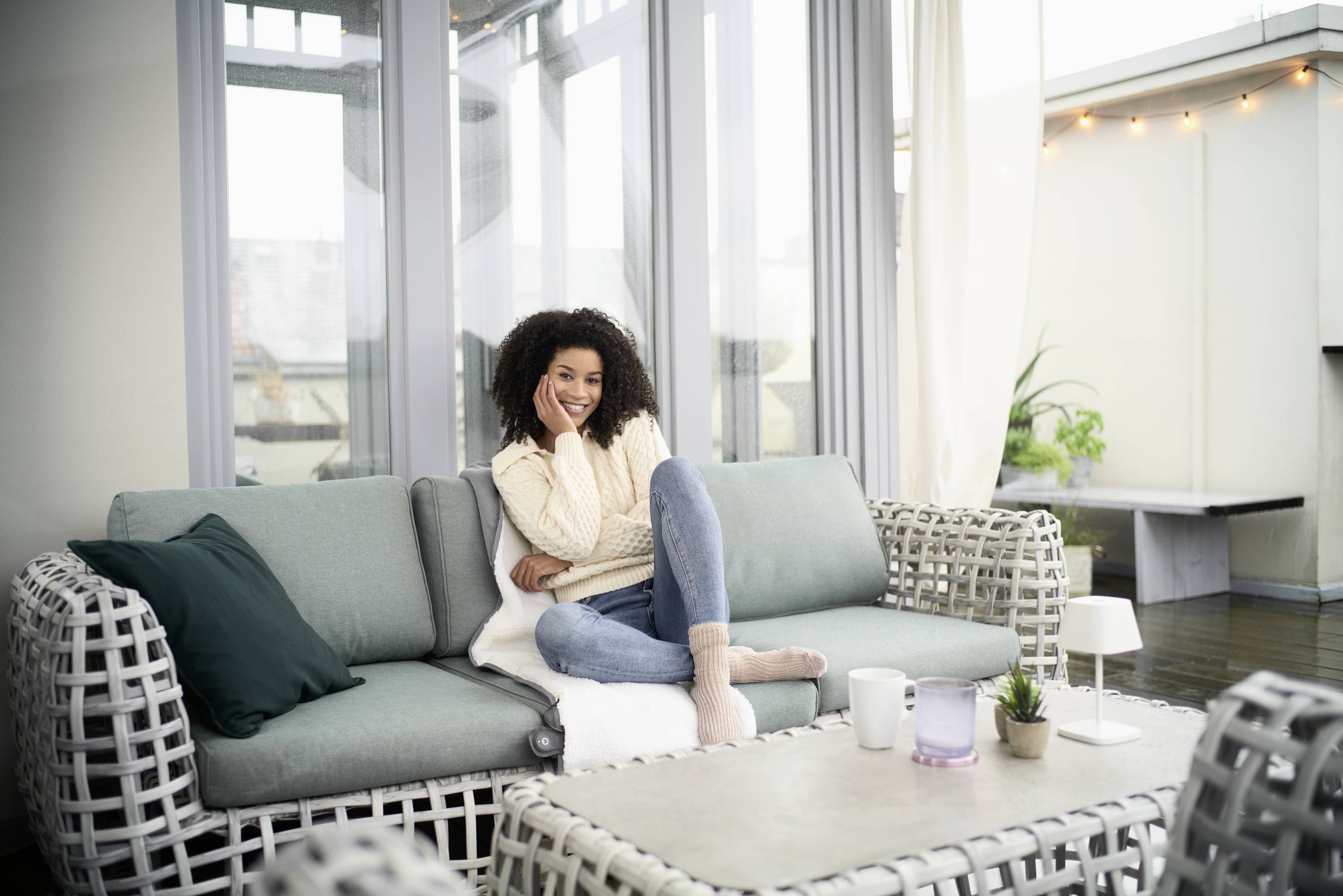 A woman sits smiling with her legs crossed on a sofa in a bright living room.
