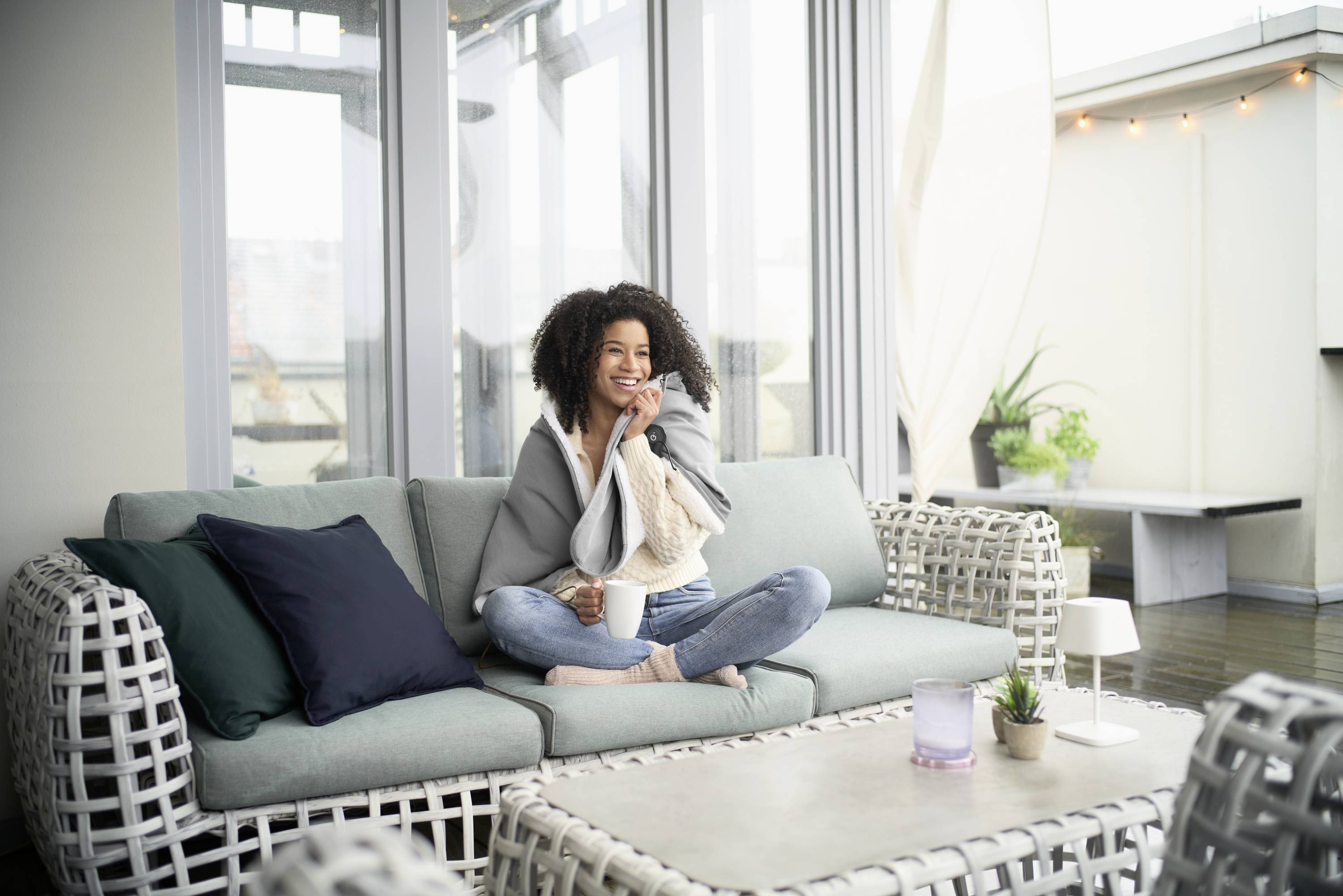 A woman sits smiling on a sofa with a coffee in a modern, bright living room with large windows and plants.