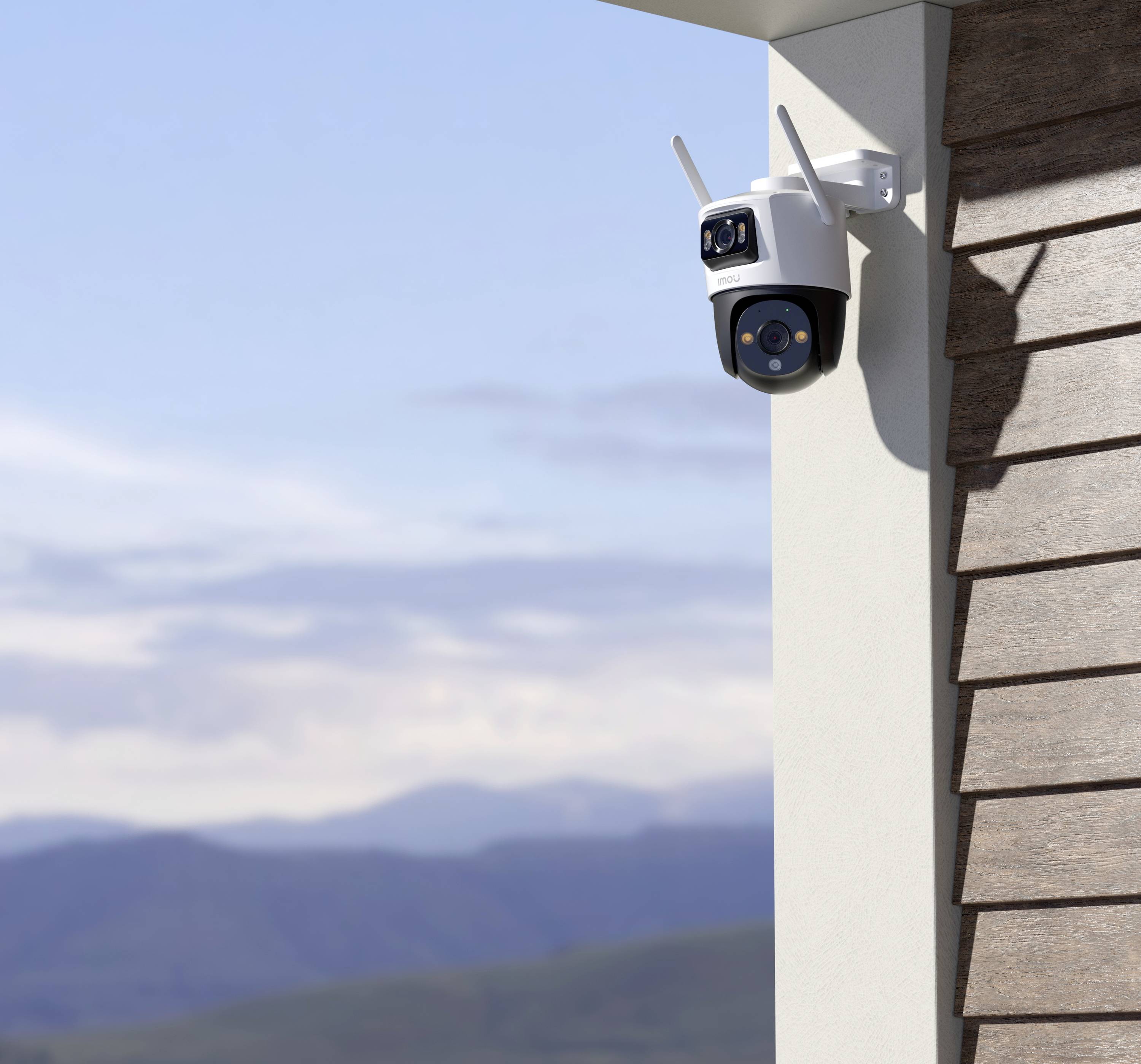 A security camera is mounted on an exterior wall, pointing towards a landscape with mountains and a blue sky in the background.
