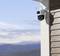 A security camera is mounted on an exterior wall, pointing towards a landscape with mountains and a blue sky in the background.