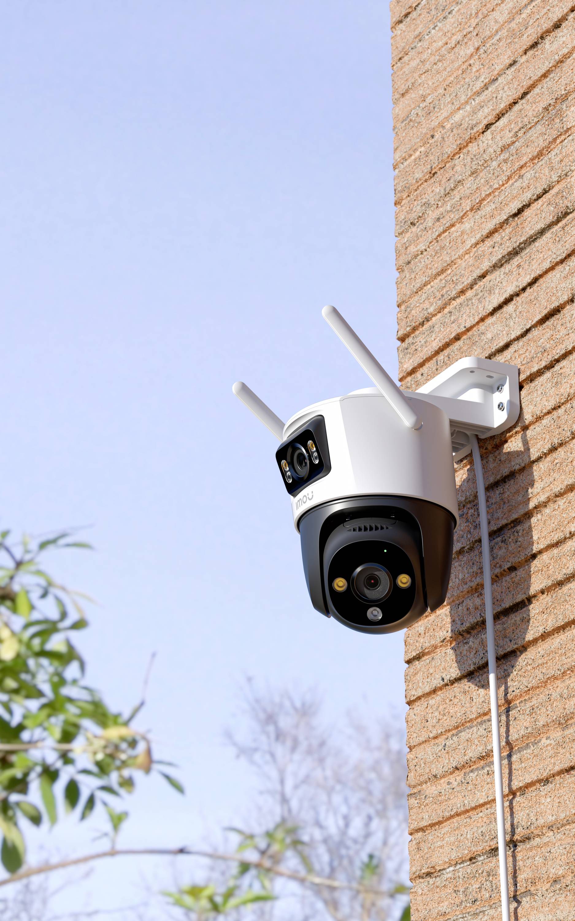 Surveillance camera mounted on a brick wall, facing outwards with a clear sky and scattered foliage in the background.