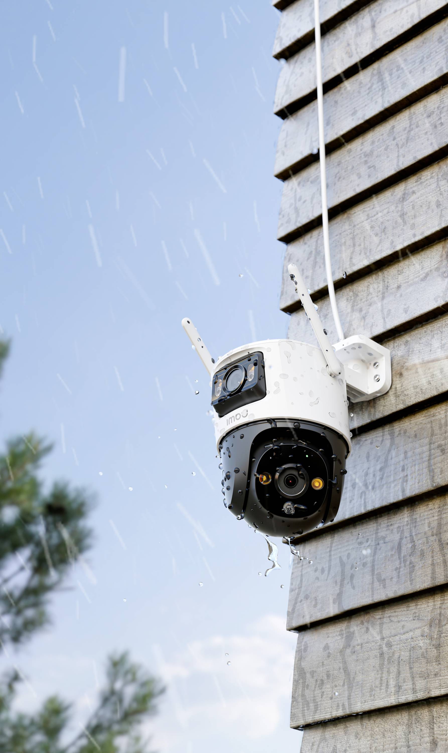 A surveillance camera is mounted on the exterior wall of a building. It is raining, and water droplets are pelting the camera.