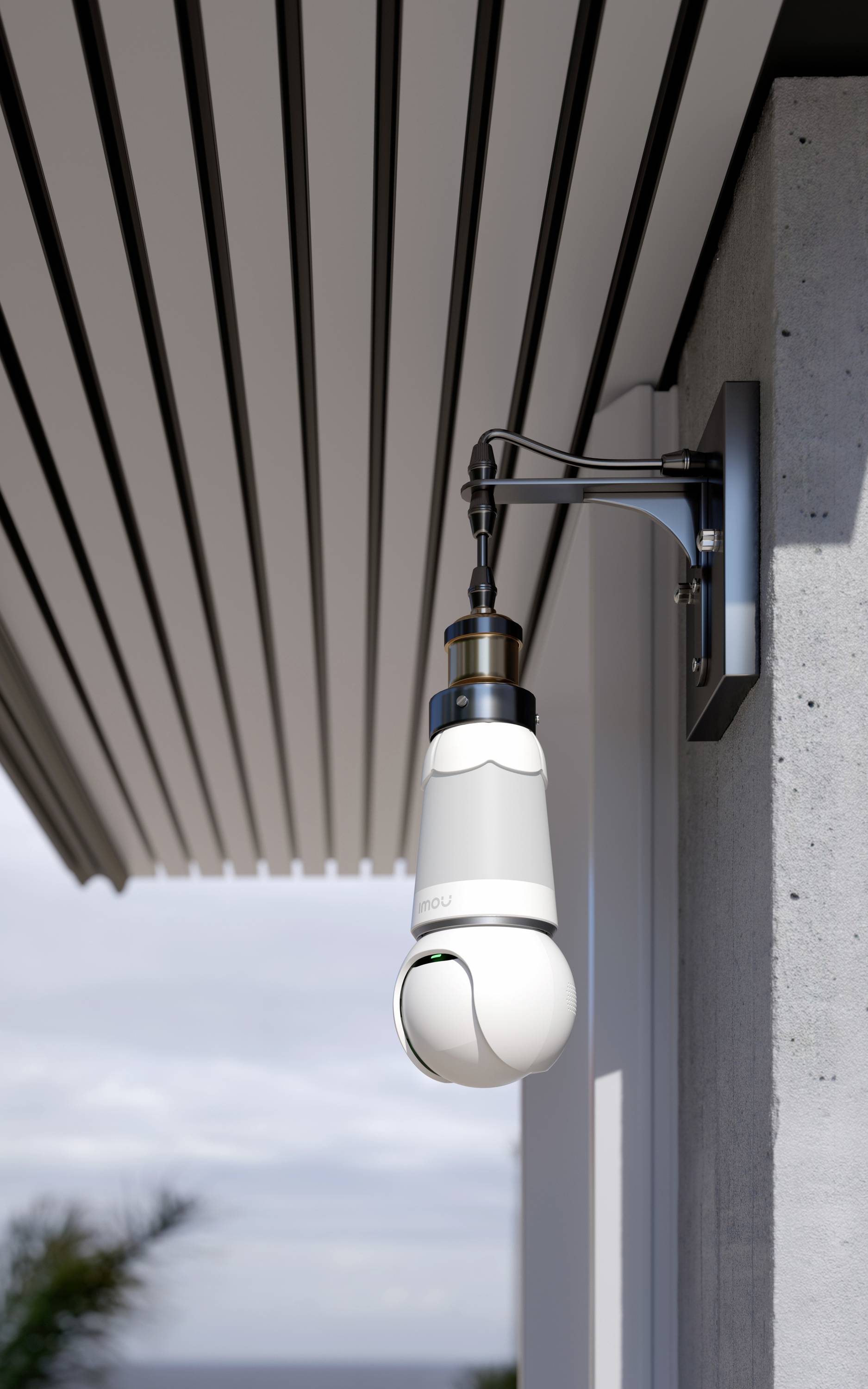 Security camera mounted on the wall of a modern building, pointing downwards, next to a covered area. Background shows sky and plants.