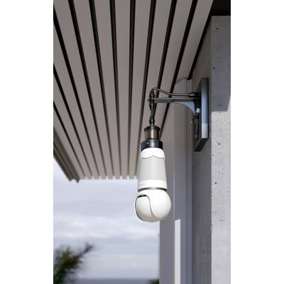 Security camera mounted on the wall of a modern building, pointing downwards, next to a covered area. Background shows sky and plants.