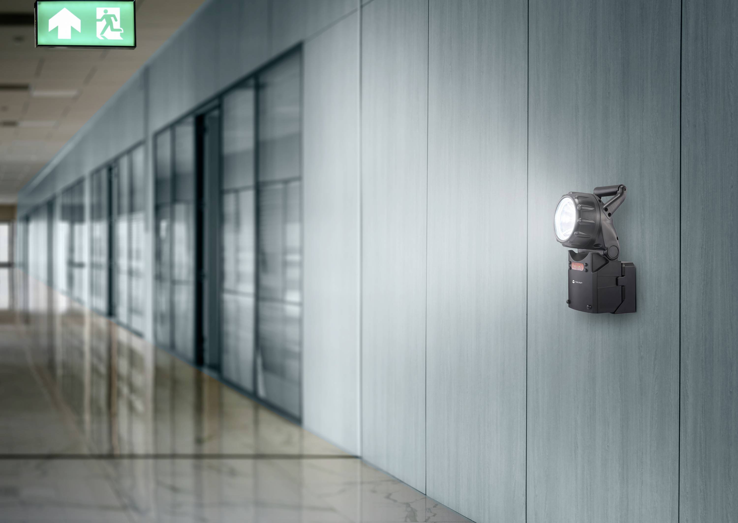 'Corridor with a green emergency exit sign on the ceiling and a wall-mounted light fitting illuminating the way.'