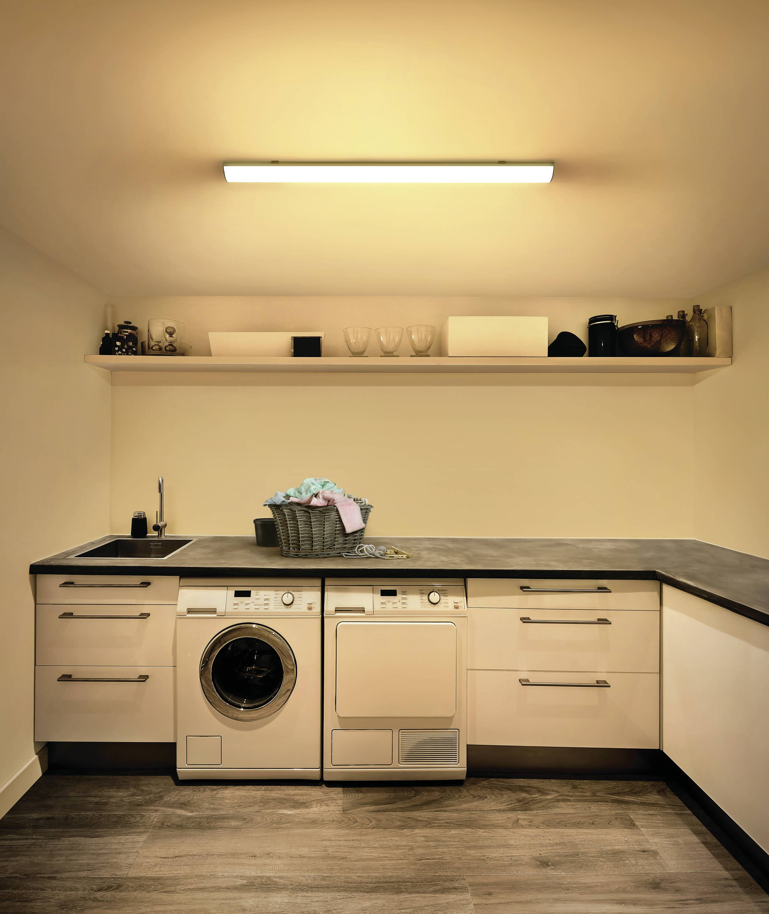 A bright utility room with a washing machine and tumble dryer underneath a worktop. A laundry basket sits on top of the counter.