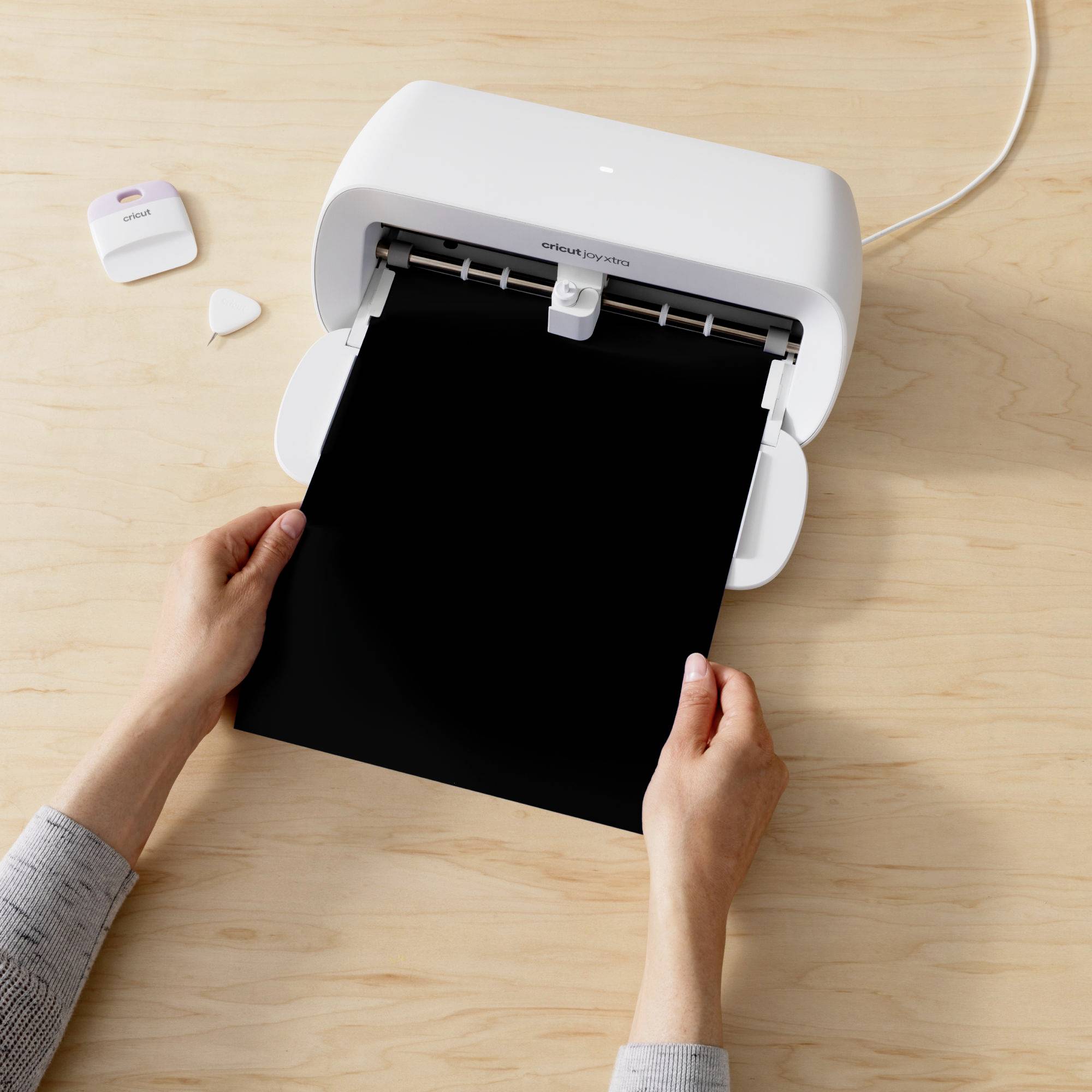 A person is cutting a black vinyl sheet with a white cutting plotter. The plotter is positioned on a wooden table.