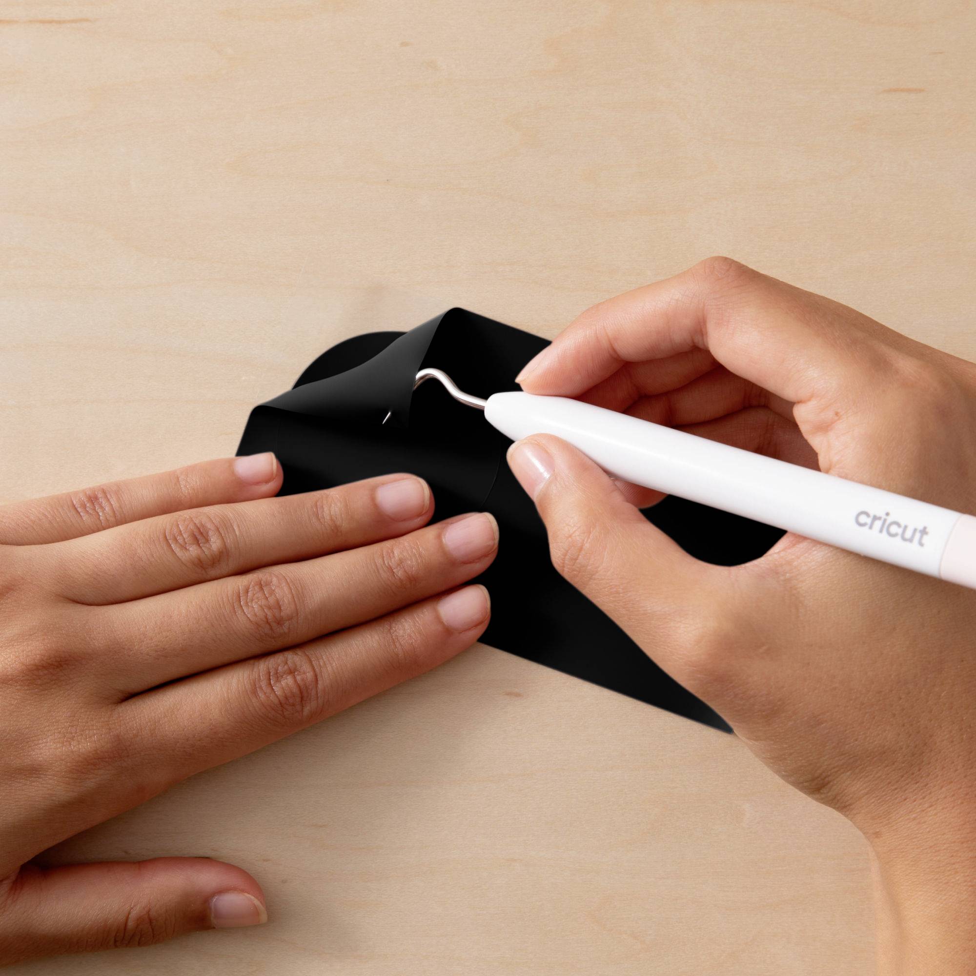 A hand holds a Cricut tool removing a black film from a wooden surface, supported by a second hand.
