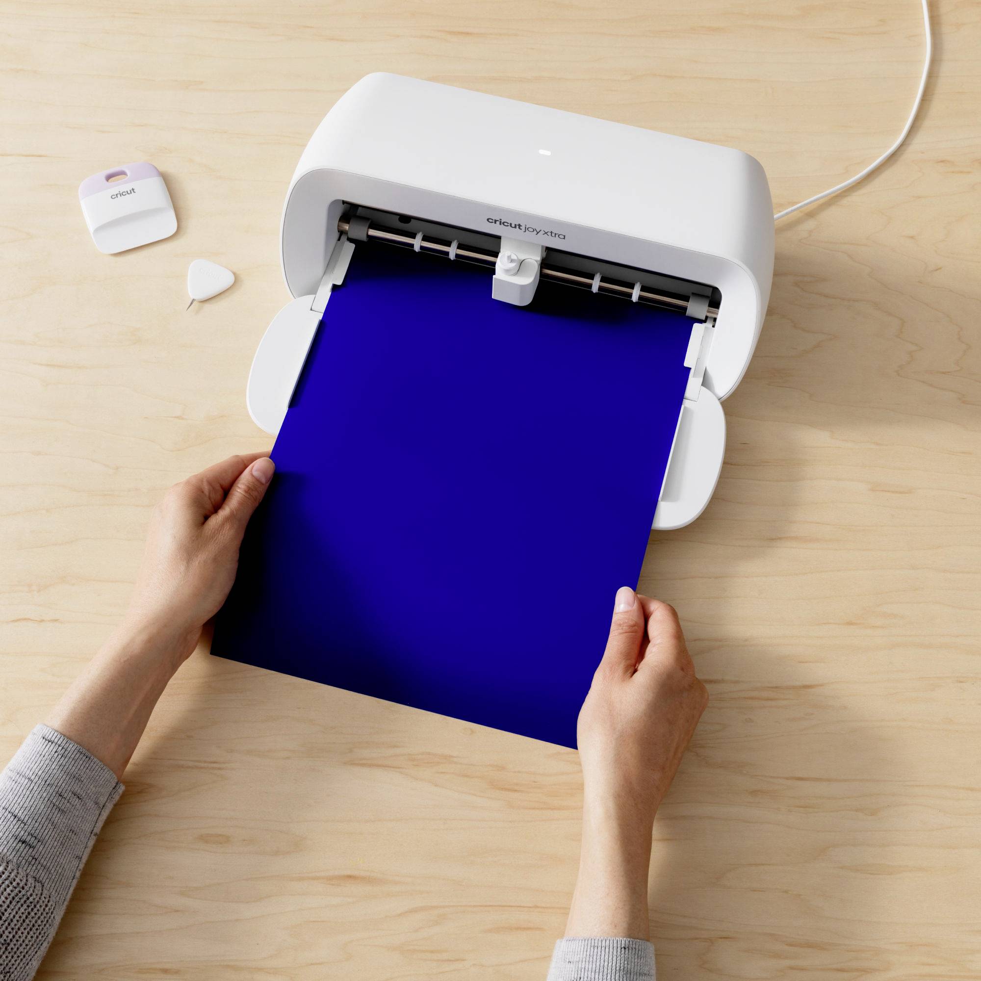 A person places a blue sheet of paper into a white cutting plotter on a table. Accessories are lying beside it.