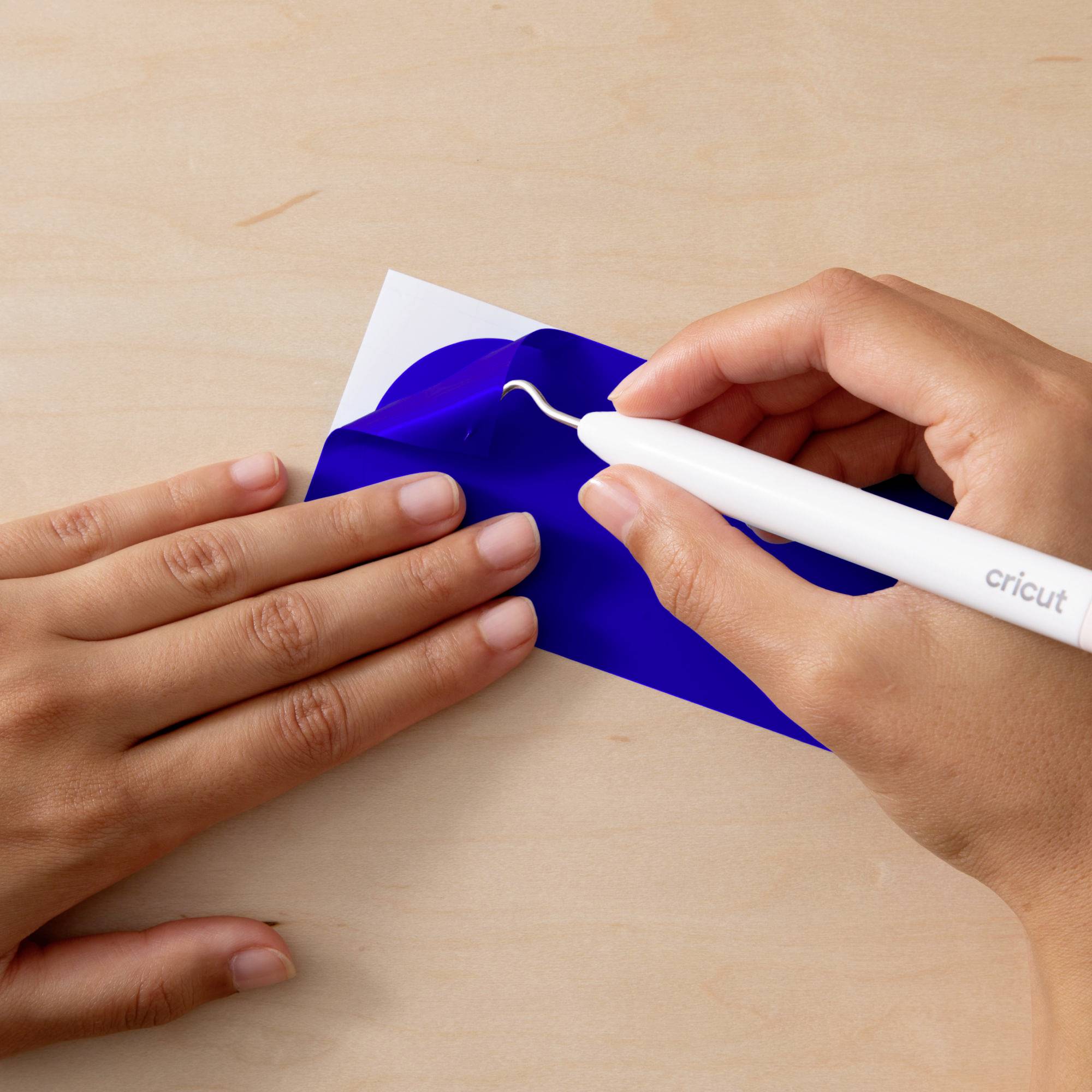 A Cricut tool is used to weed a blue vinyl design on a wooden surface. Hands are performing the work process.
