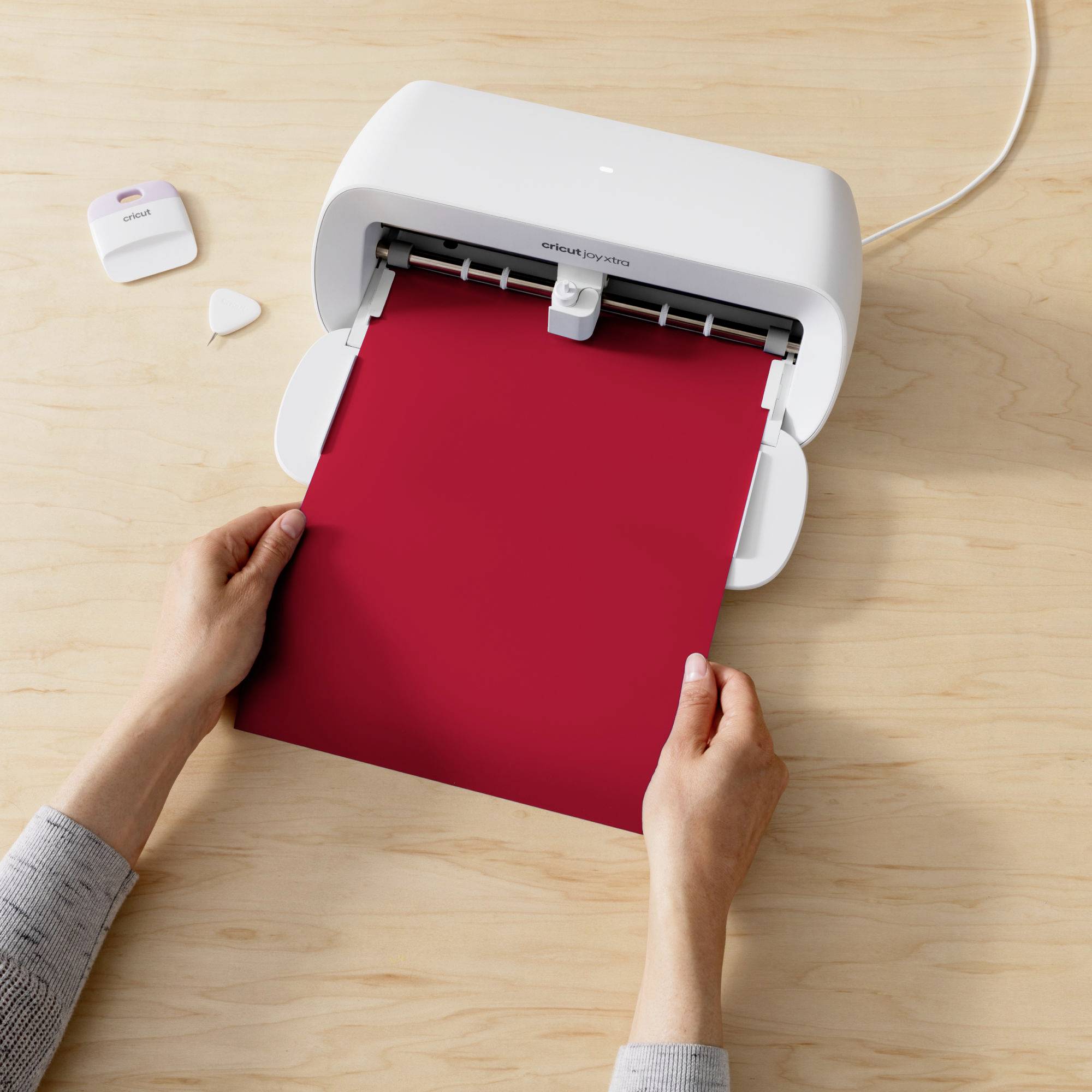 A person is holding red paper in a cutting plotter on a wooden table, ready to cut. Device in the foreground, accessories to the left.