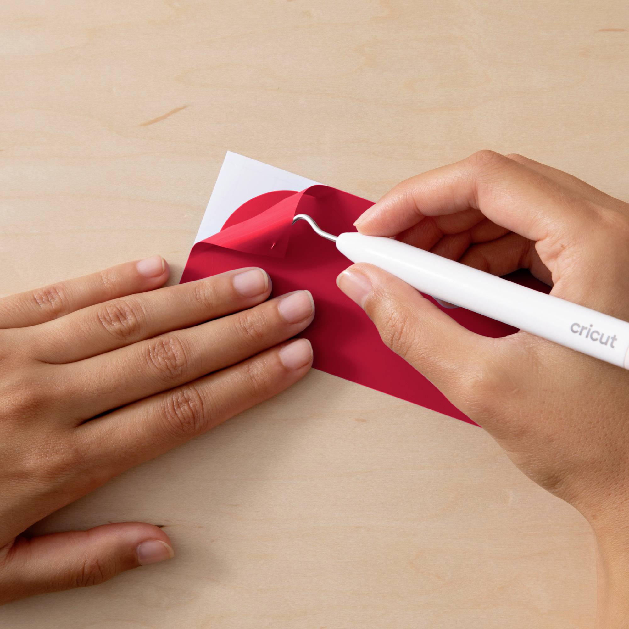 A person is precisely removing red vinyl film from a white surface on a table using a Cricut tool.