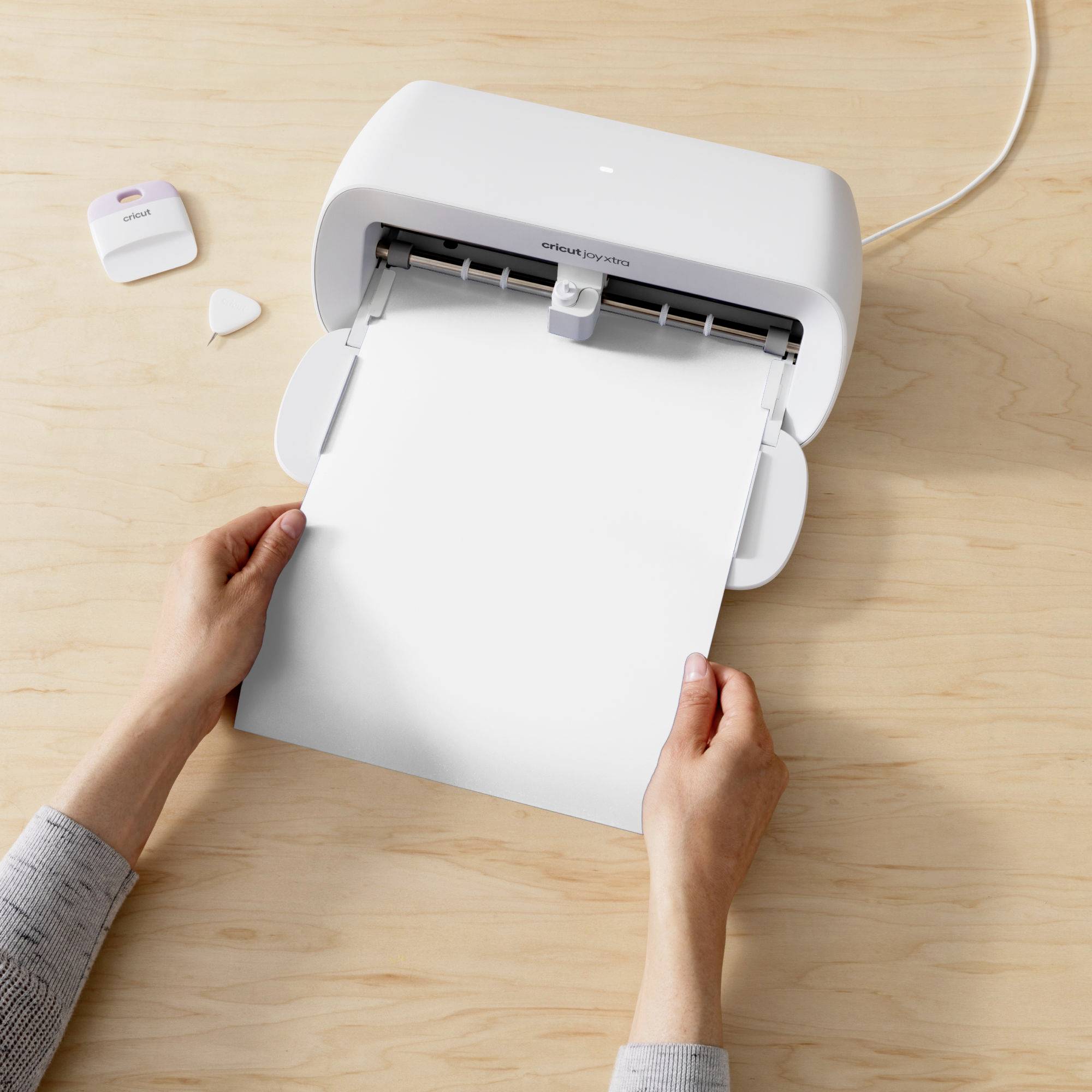 A person places a white sheet of paper into a cutting device on a wooden table. A small control panel sits nearby.