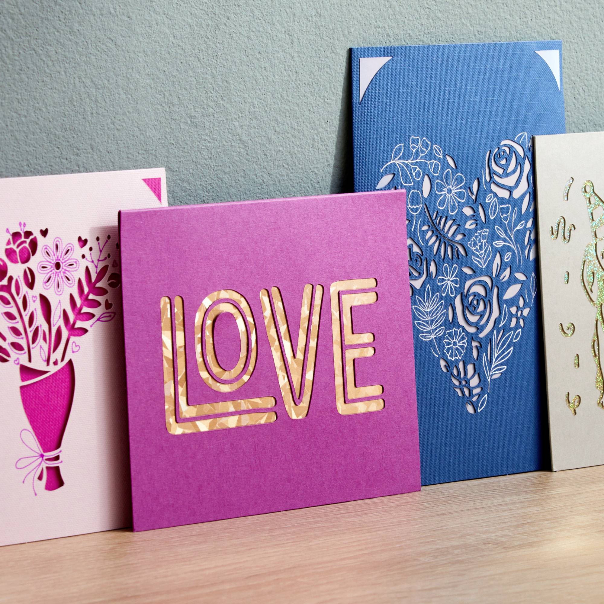 Three greetings cards with floral and heart designs displayed on a table. The middle card shows the word 'LOVE' in large letters.