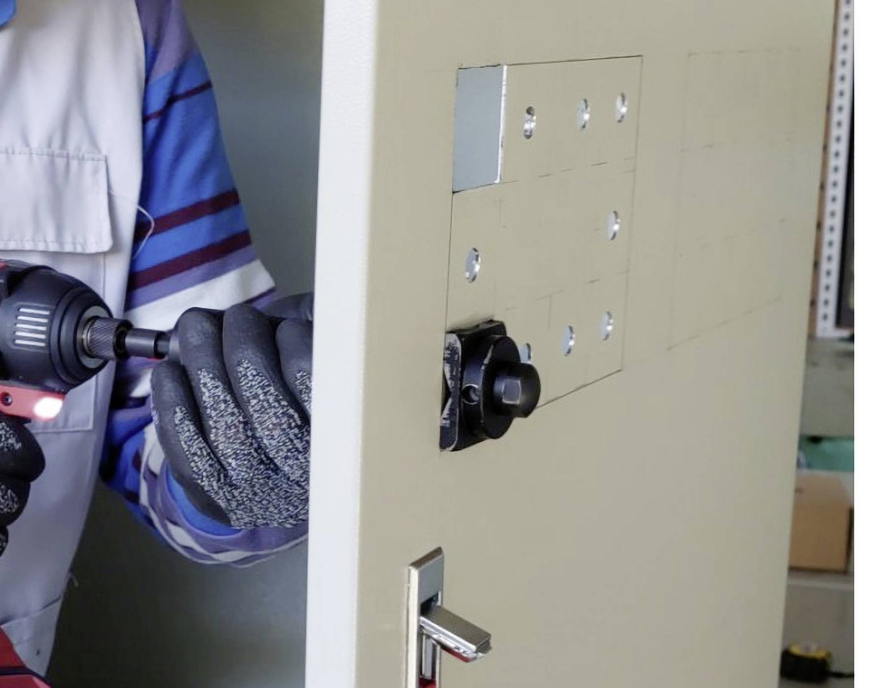 A tradesman is installing a new security lock in a metal door. He is using a cordless drill driver. He is wearing work gloves.