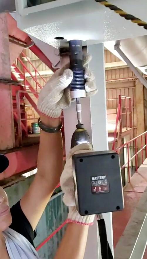 A person wearing gloves is using a cordless drill to turn a screw into a metal framework in an industrial setting.
