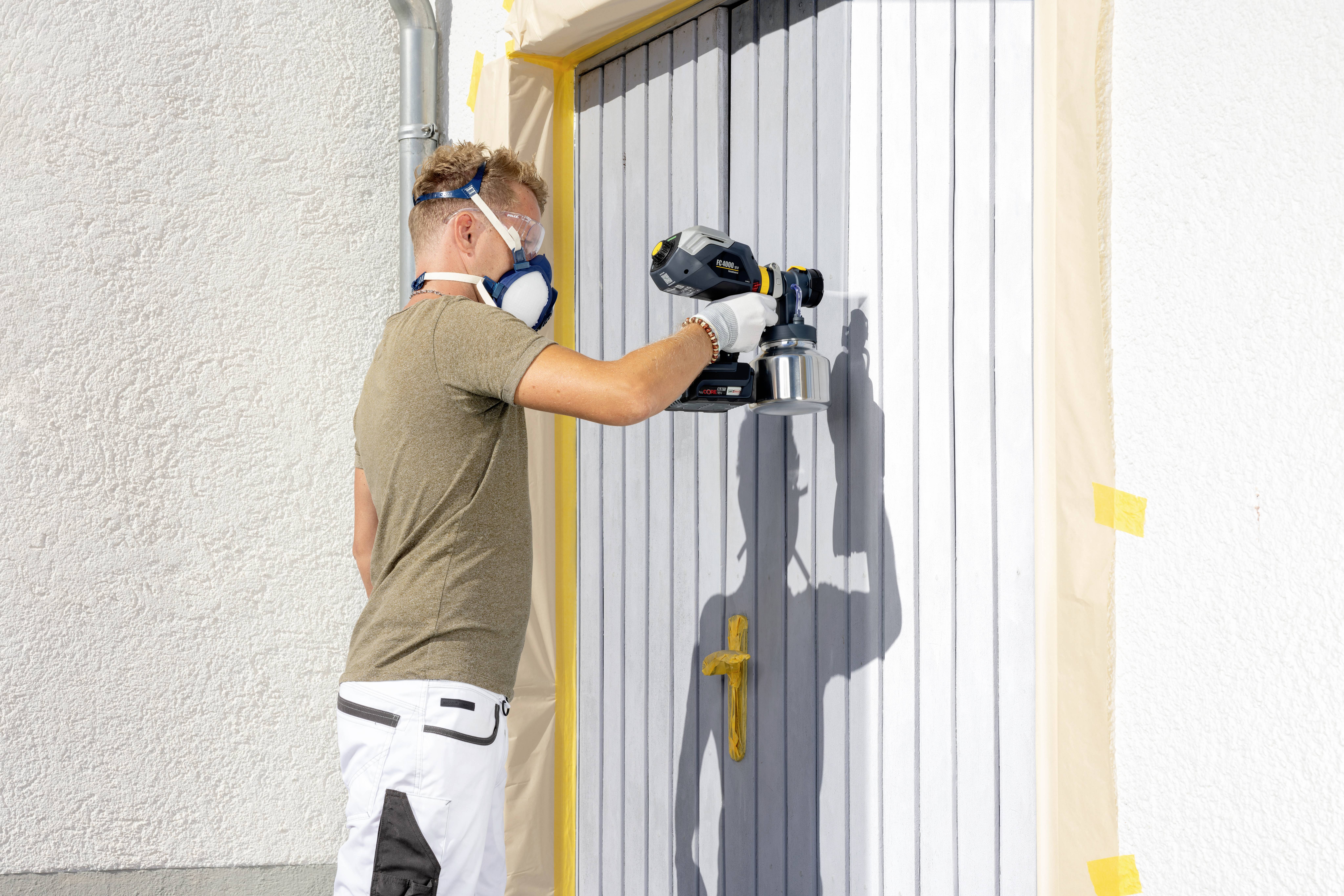 A man is spraying paint onto a door using a spray gun. The door is protected with masking film. He is wearing a respiratory mask.